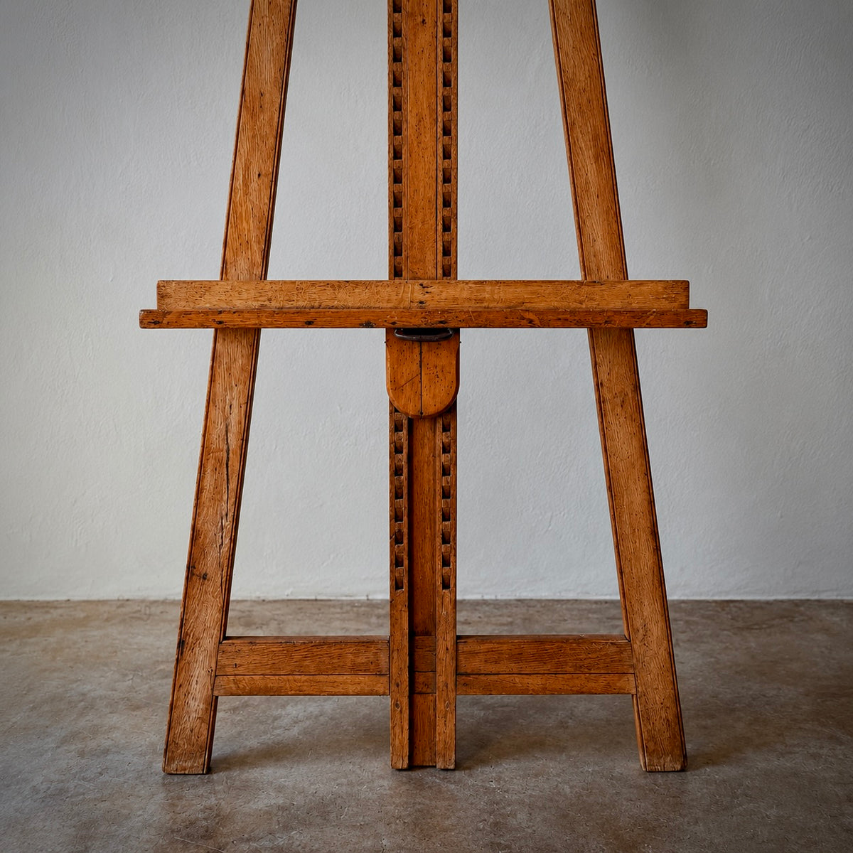 A vintage wooden easel with a warm medium-brown finish, standing on a concrete floor against a neutral off-white wall.