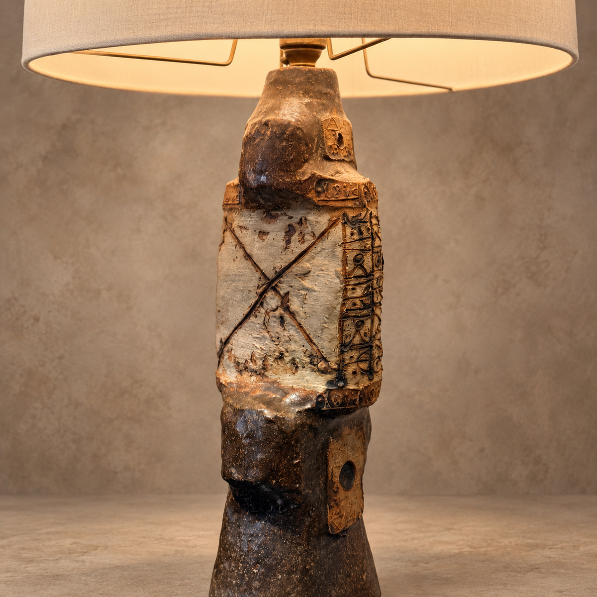 A sculptural table lamp with a textured ceramic warrior figure base and a beige drum-style shade, shown lit against a neutral textured studio background.