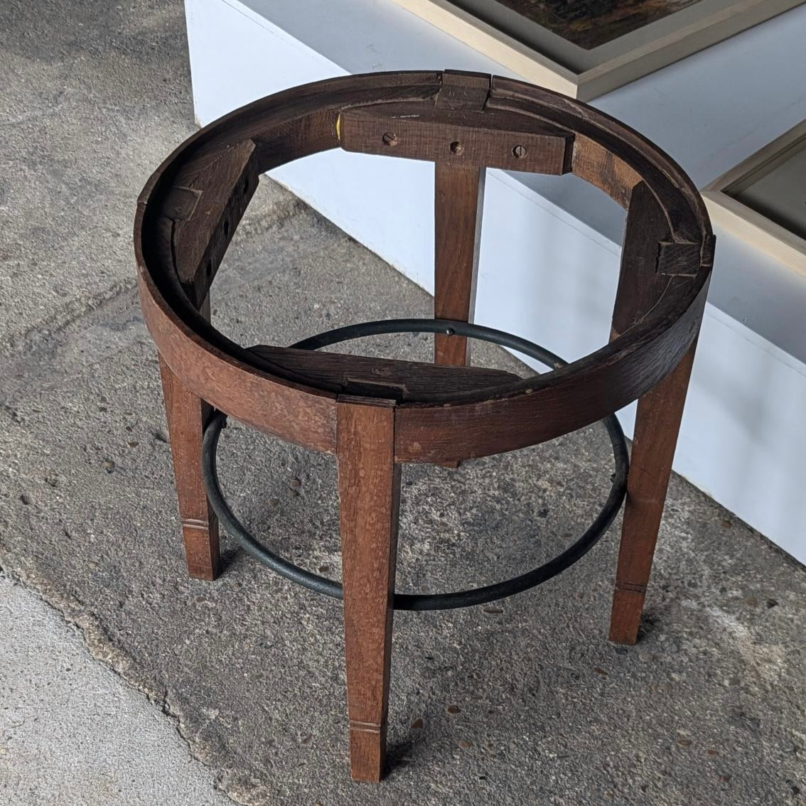 A rustic dark brown wooden stool base or table frame with four legs and a circular black metal ring base, photographed on a concrete floor.