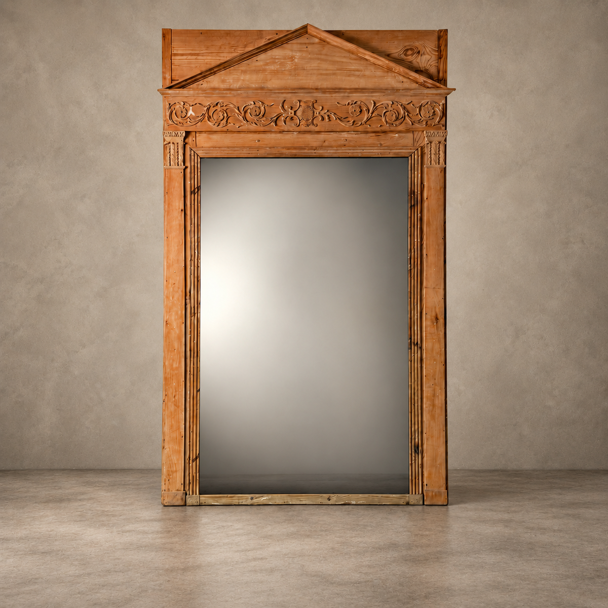 A large rectangular wall mirror with an ornate carved wooden frame and a triangular pediment top, shown standing against a textured gray wall and concrete floor.