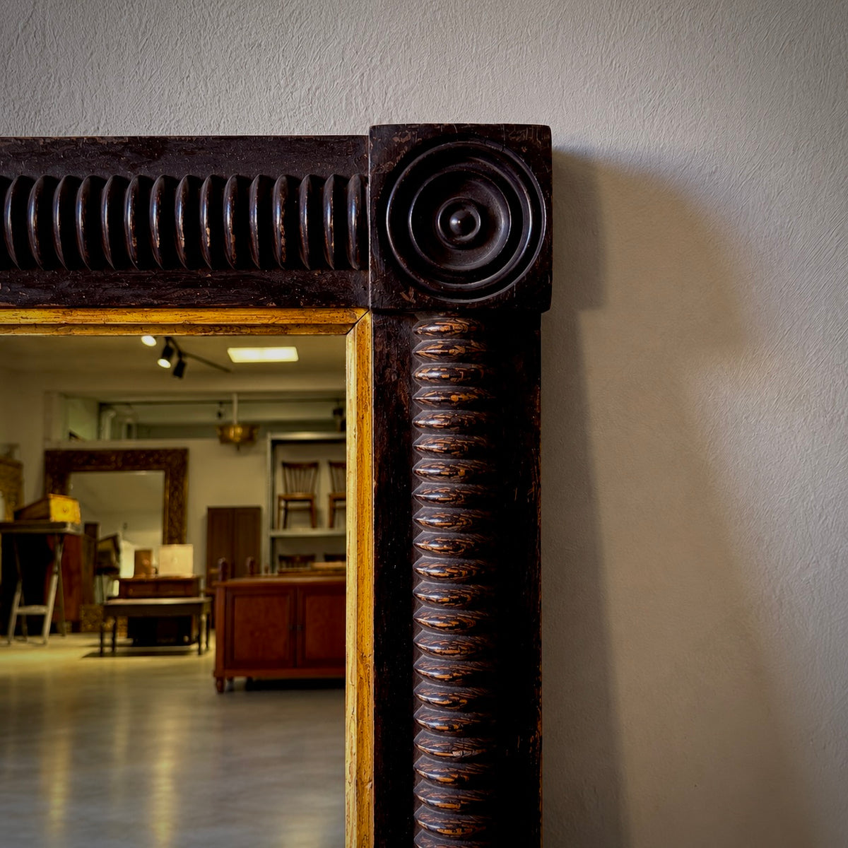 Close-up of an ornate dark brown carved wooden mirror frame with a spiral column detail and a gold-toned inner border, set against a light gray wall with a showroom interior visible in the mirror reflection.