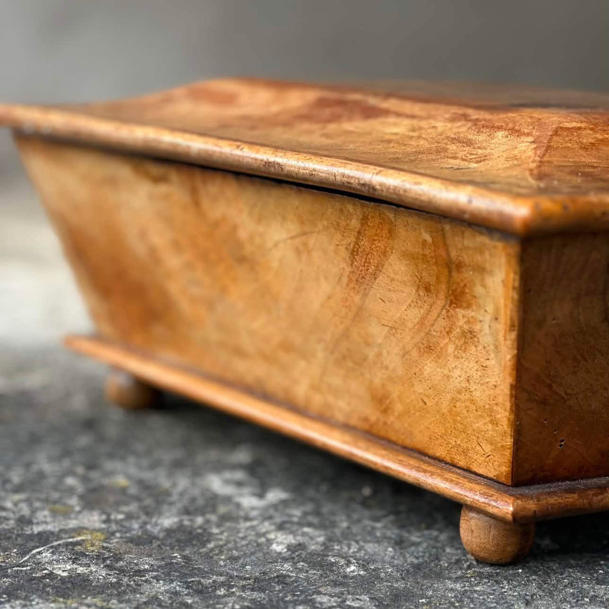 A close-up of a rectangular wooden box with a hinged lid, resting on a textured gray stone surface.