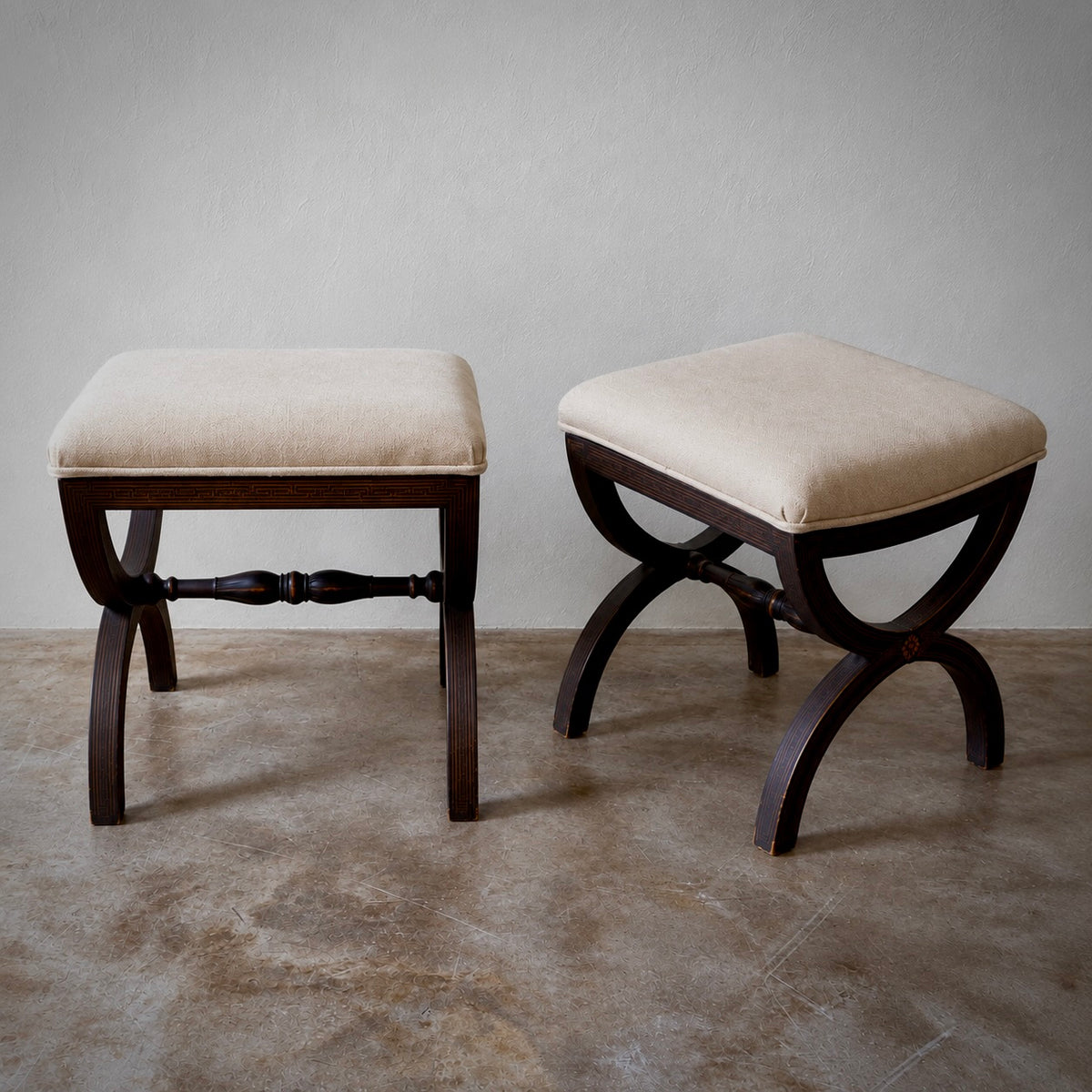 Two wooden bench stools with cream upholstered square seats and curved dark wood legs, shown on a concrete floor against a neutral gray wall.