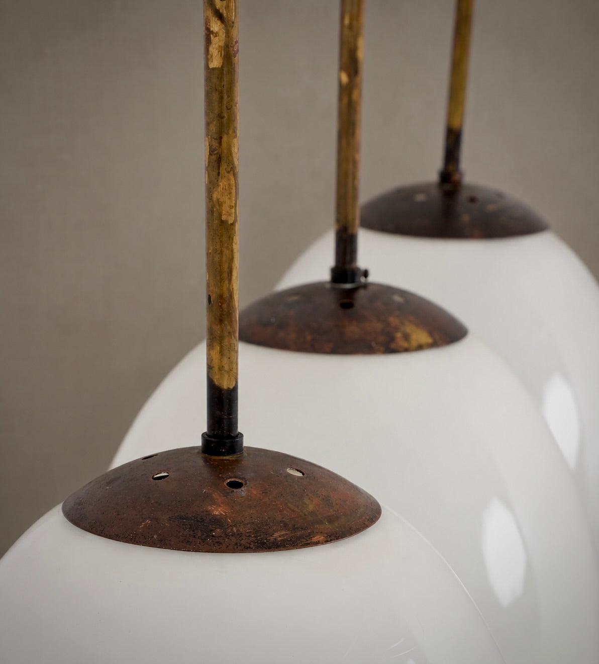 A close-up of a modern pendant light fixture with glossy white glass globes, rusted bronze metal caps, and vertical brass-colored rods against a neutral gray background.