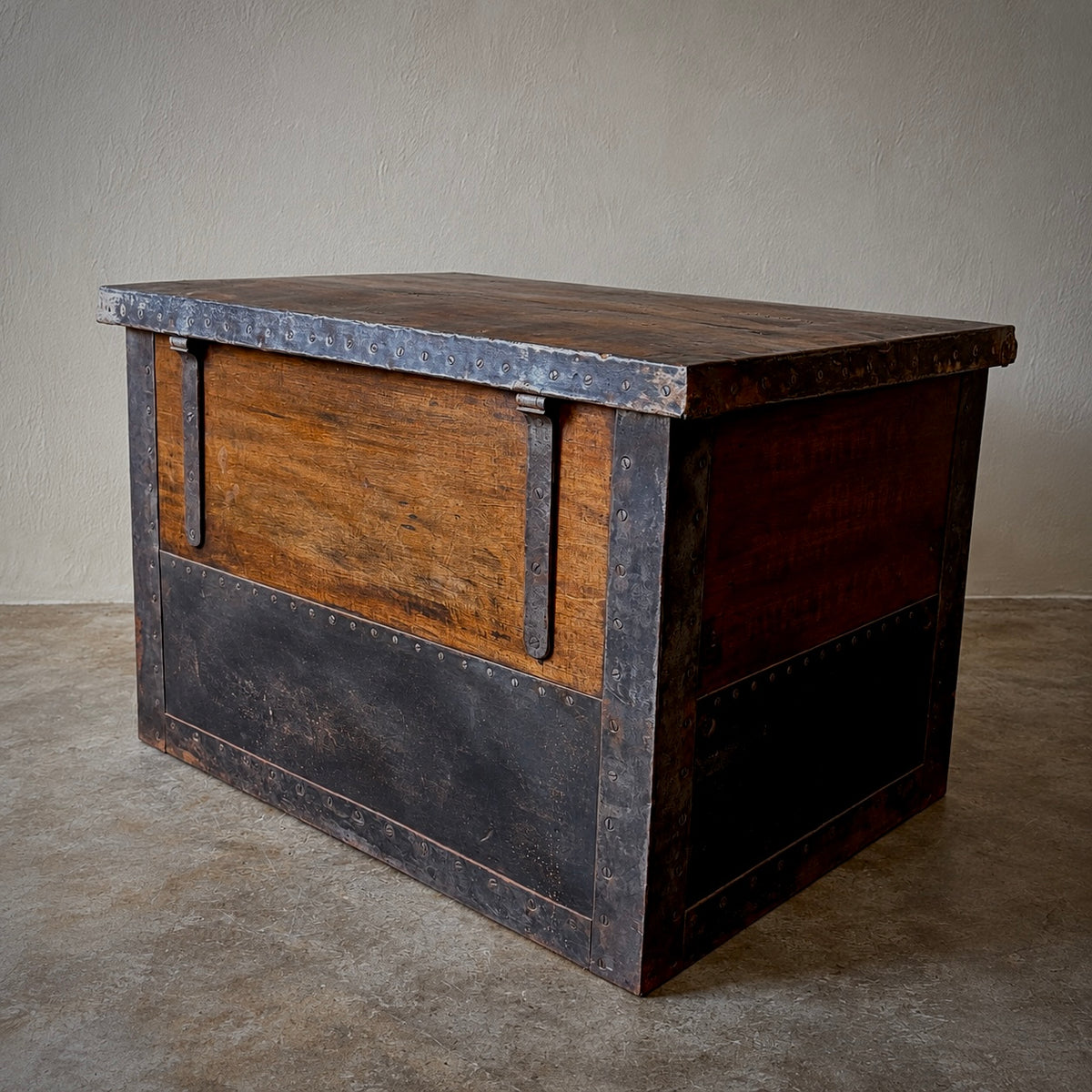 A rustic industrial-style wooden chest or trunk with dark metal framing, rivets, and a weathered wood grain finish, photographed indoors against a plain wall and concrete floor.
