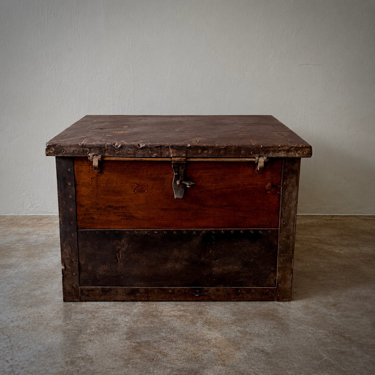 A vintage wooden storage chest with a weathered dark brown leather top, a warm medium-brown wooden front panel, and a darker lower section, photographed against a plain light gray wall and concrete floor.
