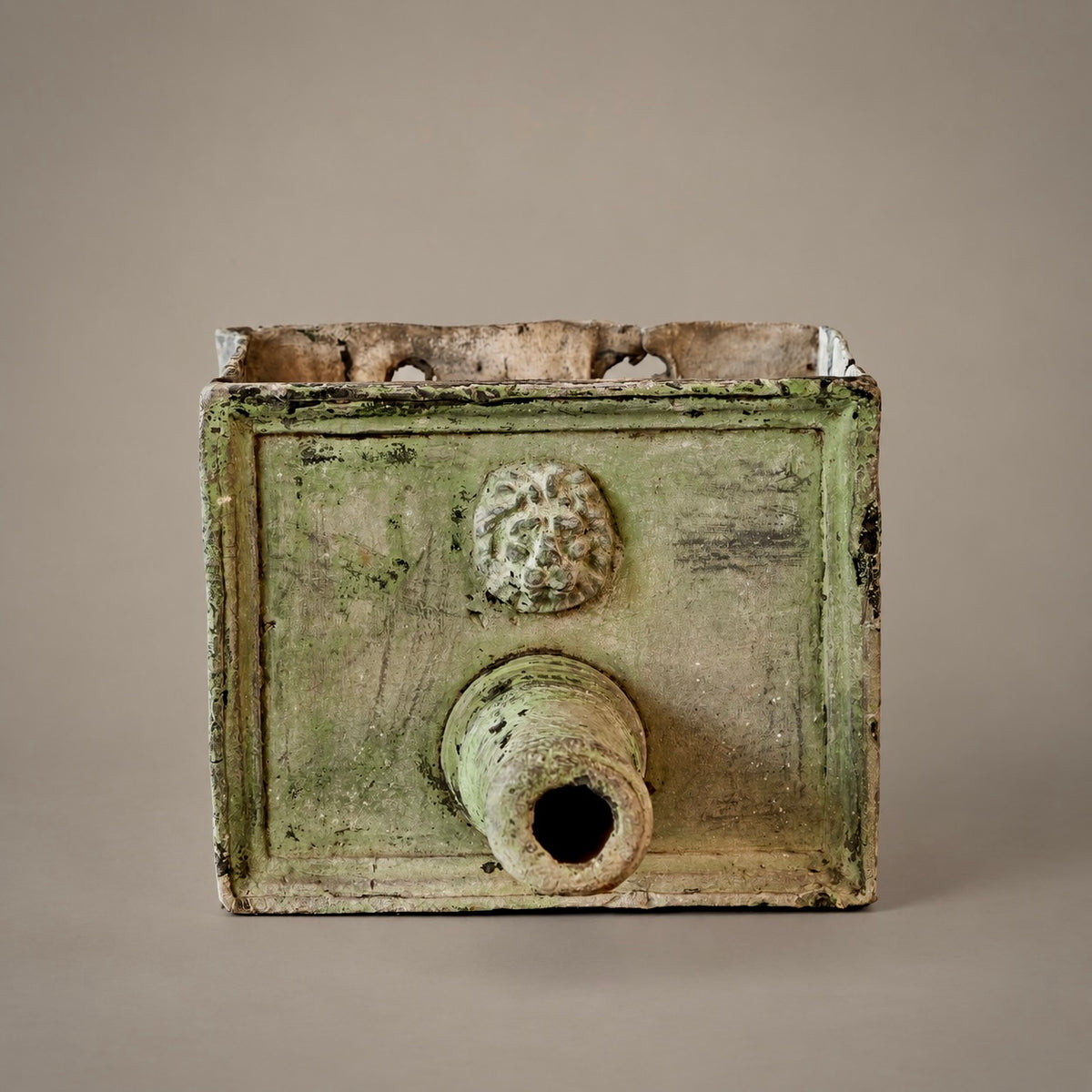 An ancient rectangular metal fountain head with a protruding cylindrical opening and a sculpted lion head motif on the front, shown against a neutral gray background.