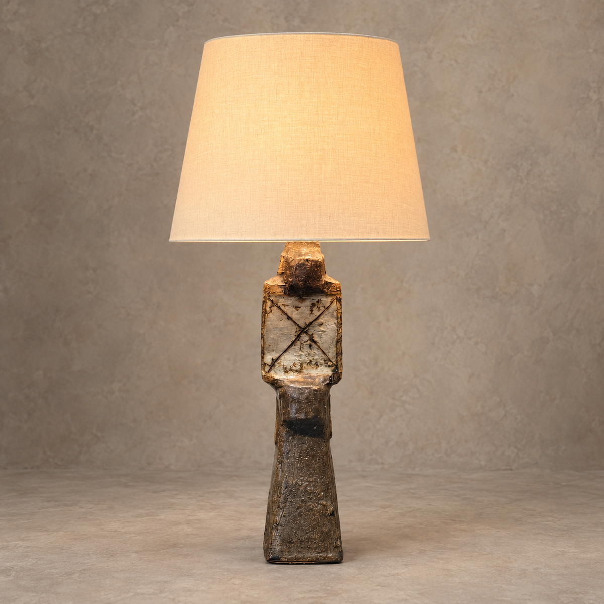 A sculptural table lamp with a textured, rustic dark brown ceramic figure base and a warm beige fabric drum shade, set against a neutral textured gray background.