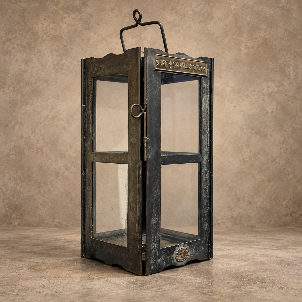 A rustic black metal lantern with a distressed finish, featuring clear glass panels, a top hanging loop, and decorative brass-toned hardware, set against a textured beige studio backdrop.