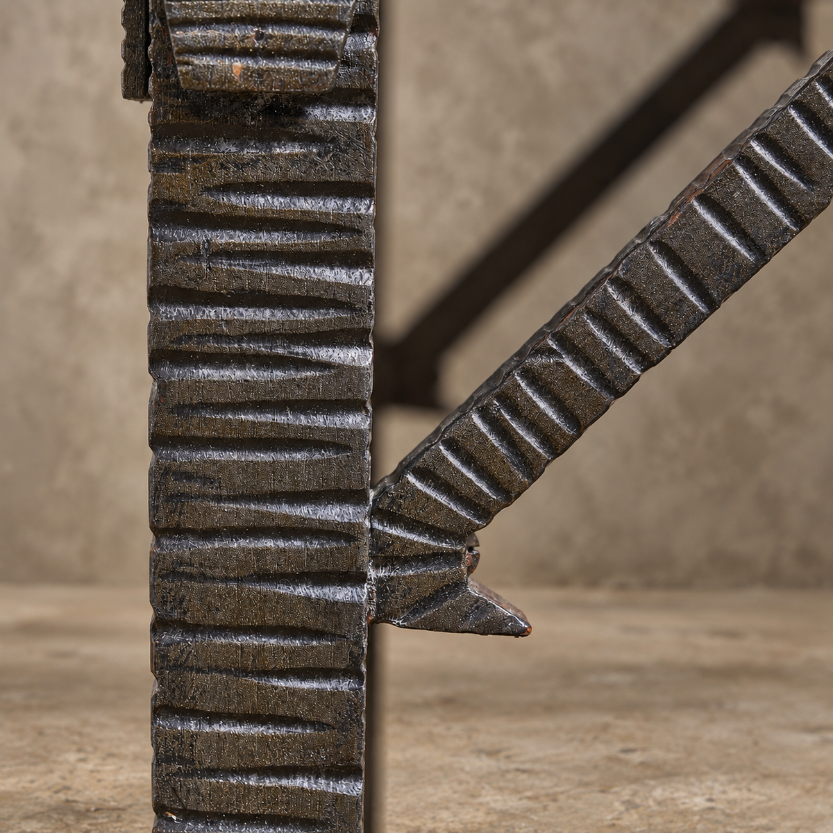 Close-up detail of a dark metal table leg with a ribbed vertical post and a diagonal support brace, shown against a warm beige textured background.