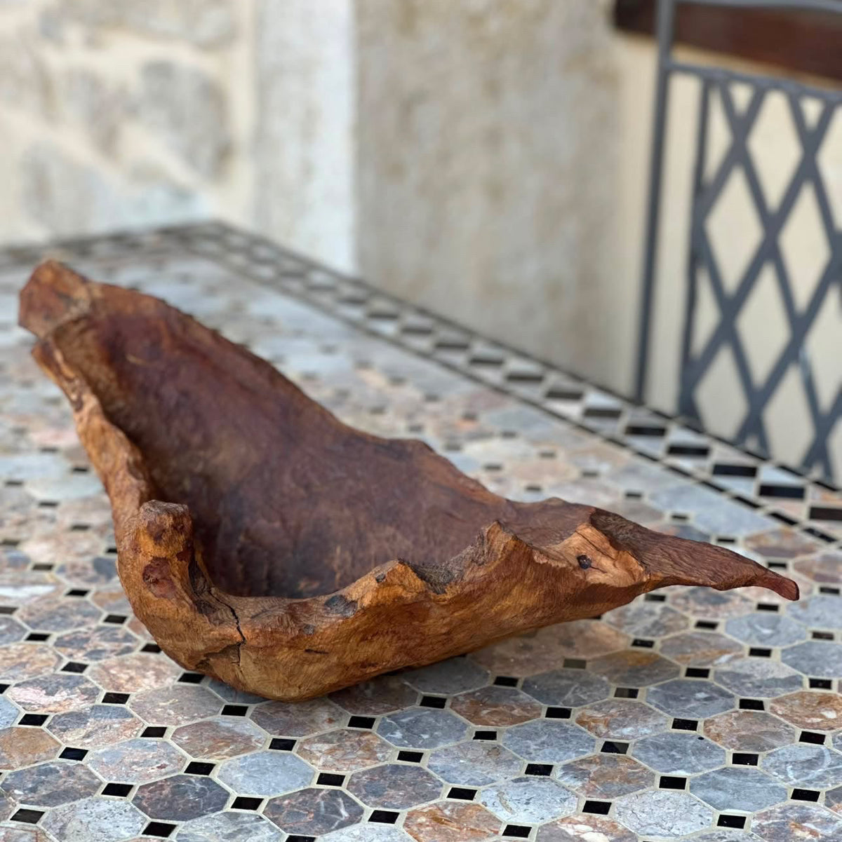 A rustic, hand-carved wooden bowl with natural bark edges, displayed on a mosaic tile patio table in front of a stone wall.