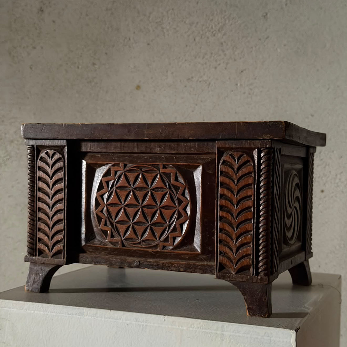 An antique dark brown carved wooden chest with intricate geometric and leaf patterns, displayed on a white pedestal against a textured beige wall.