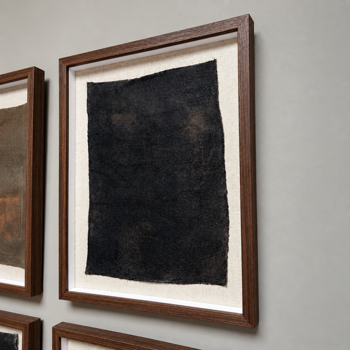 A framed abstract art piece featuring a textured black rectangular sheet mounted on a cream mat, housed in a dark brown wooden frame, displayed on a neutral gray wall.