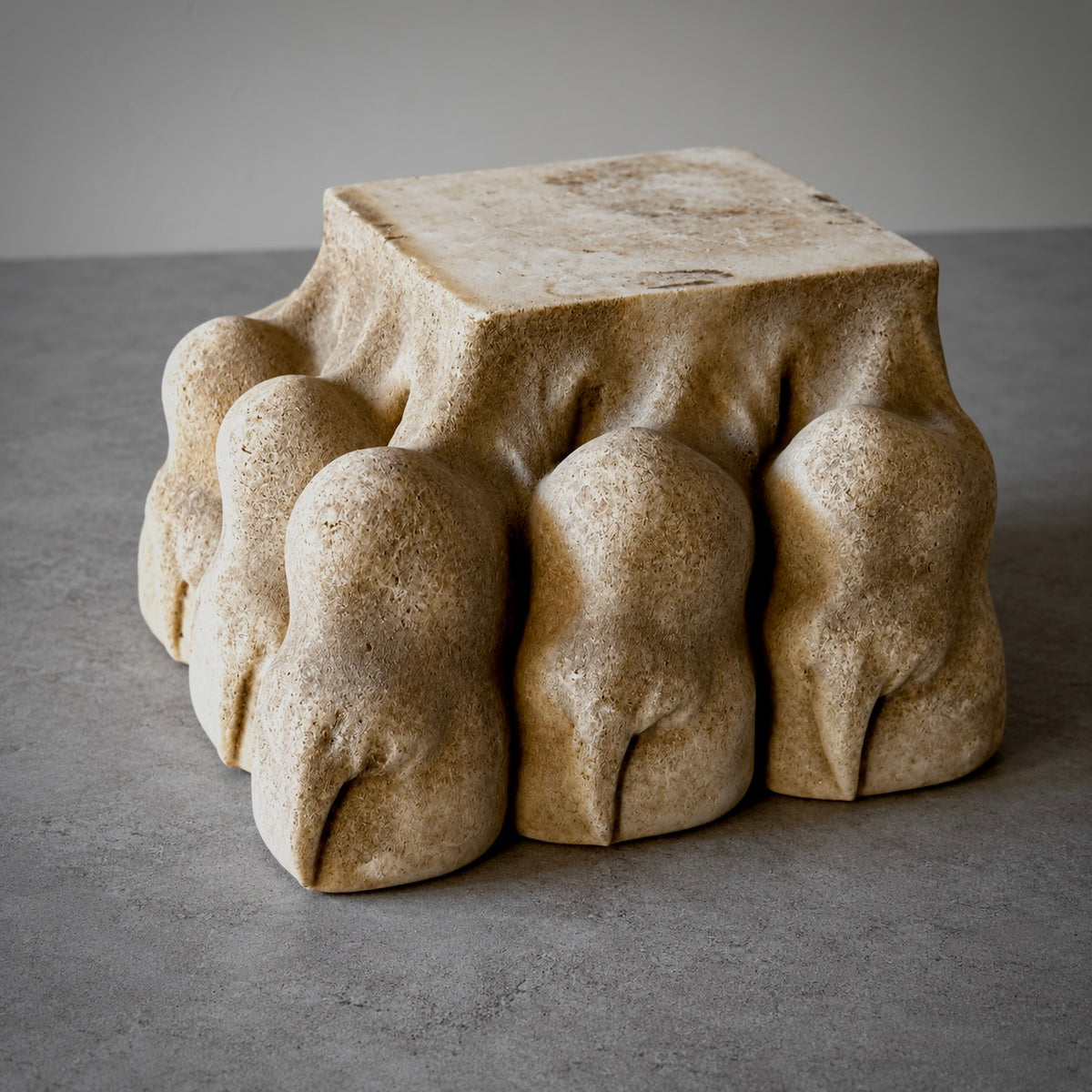 A sculptural stone lions paw made from a beige travertine-like block, supported by five rounded, claw-like paws. The object is photographed on a neutral gray surface against a matching gray background.