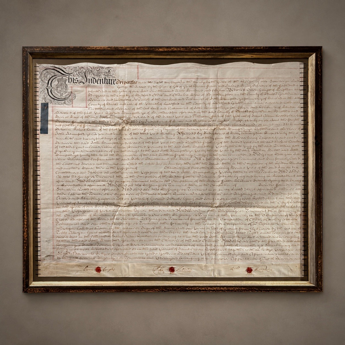 A framed property deed, featuring the original parchment document inside a large, ornate dark brown wooden frame., mounted in a dark wood frame against a neutral gray wall.