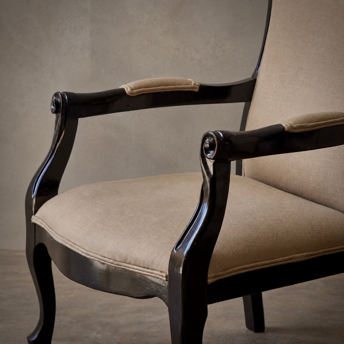 A close-up of a classic armchair with a glossy black carved wooden frame and beige upholstered seat and back cushions, set against a neutral textured gray background.