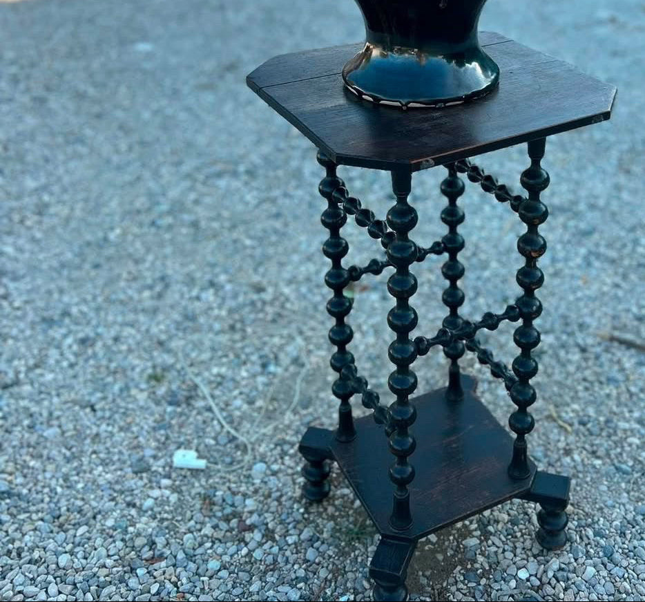 A decorative ceramic vase with a dark green and black glaze sits on a small dark wooden pedestal table outdoors on gravel, with shipping containers in the background. White text overlays show the dimensions and price of the item.