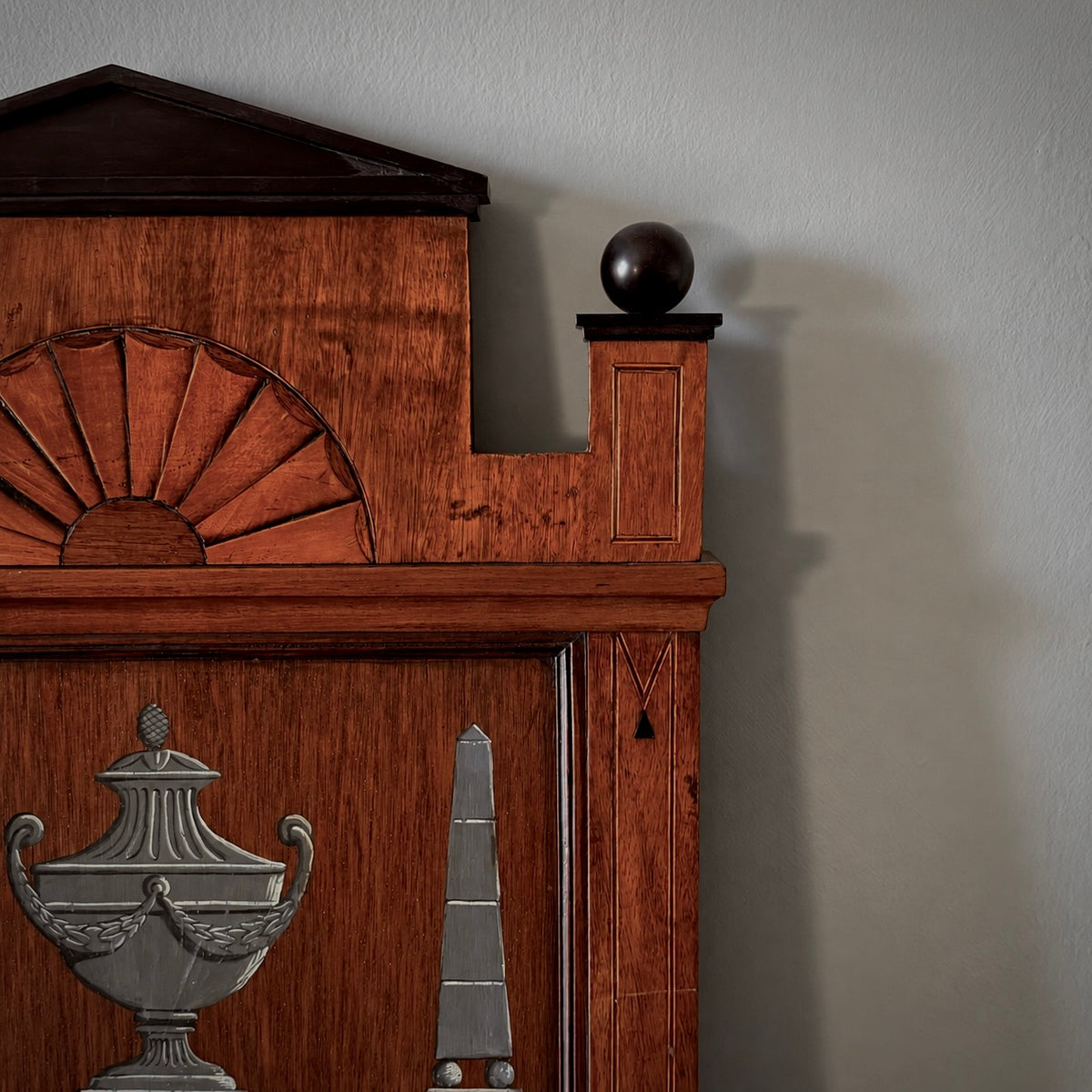Close-up of a dark reddish-brown wooden headboard with carved decorative details, including a fan motif and sculptural metal accents, set against a plain light gray wall.
