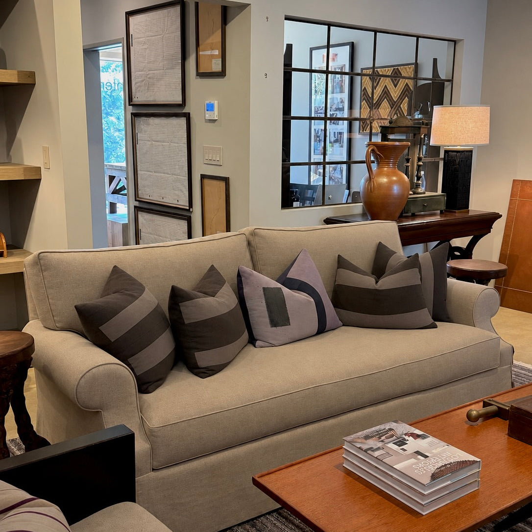 A beige upholstered sofa in a styled living room setting, decorated with multiple dark gray and striped throw pillows, positioned next to a wooden coffee table and shelves in the background.