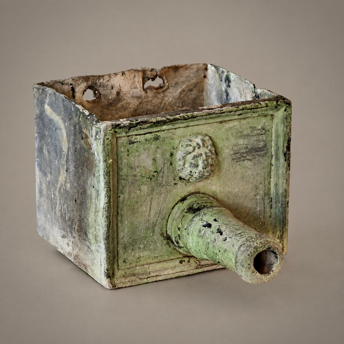 An ancient rectangular metal fountain head with a protruding cylindrical opening and a sculpted lion head motif on the front, shown against a neutral gray background.