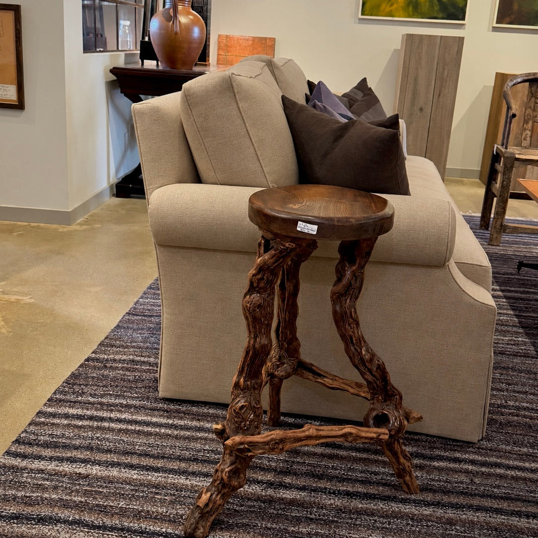 A rustic wooden stool with gnarled, branch-like legs stands in front of a beige upholstered sofa in a furniture showroom. The stool has a round dark wood seat and is positioned on a striped gray rug.