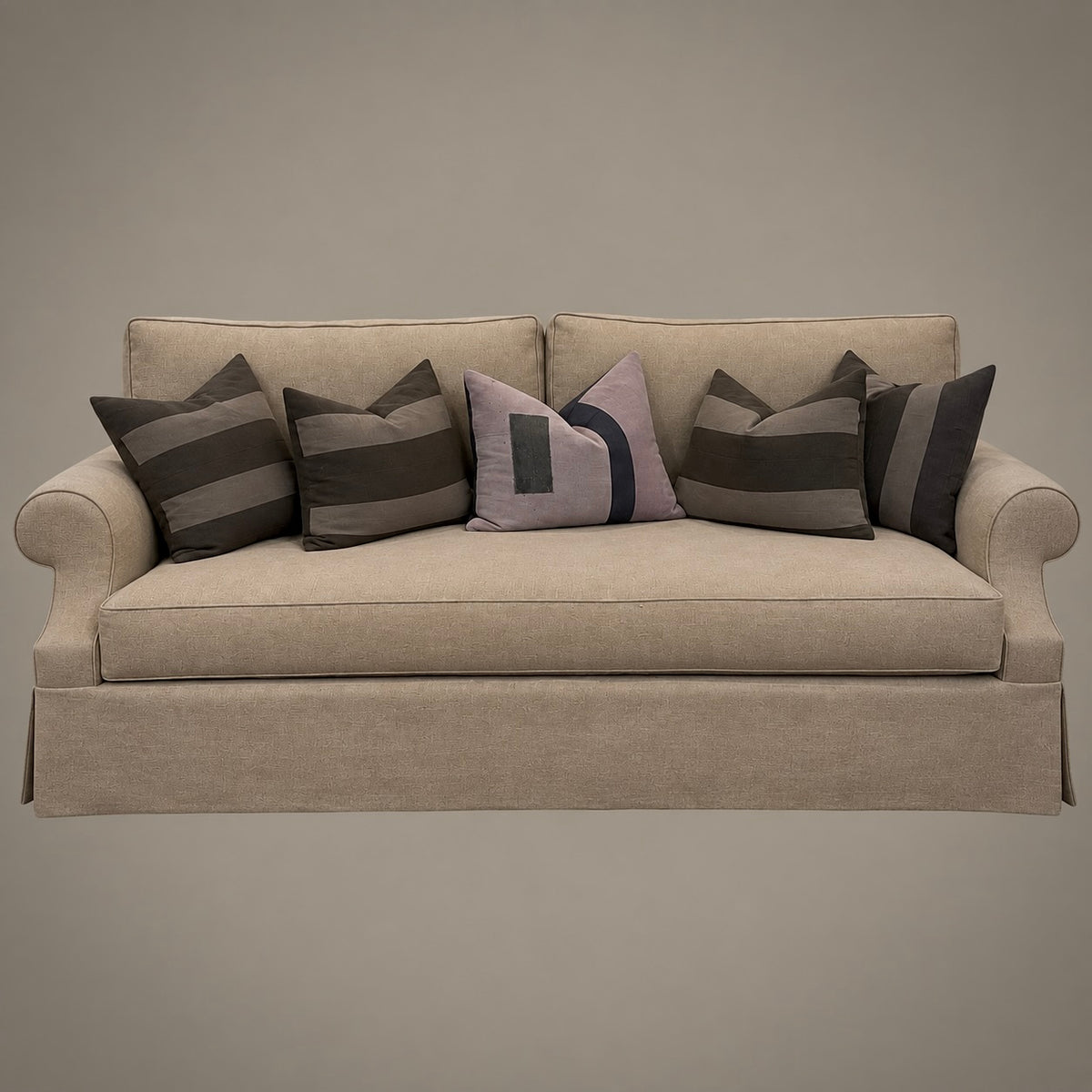 A beige upholstered sofa with rolled arms and a skirted base, shown against a plain taupe background. The sofa is styled with multiple throw pillows in dark gray, charcoal, and a muted lavender-gray patterned design.