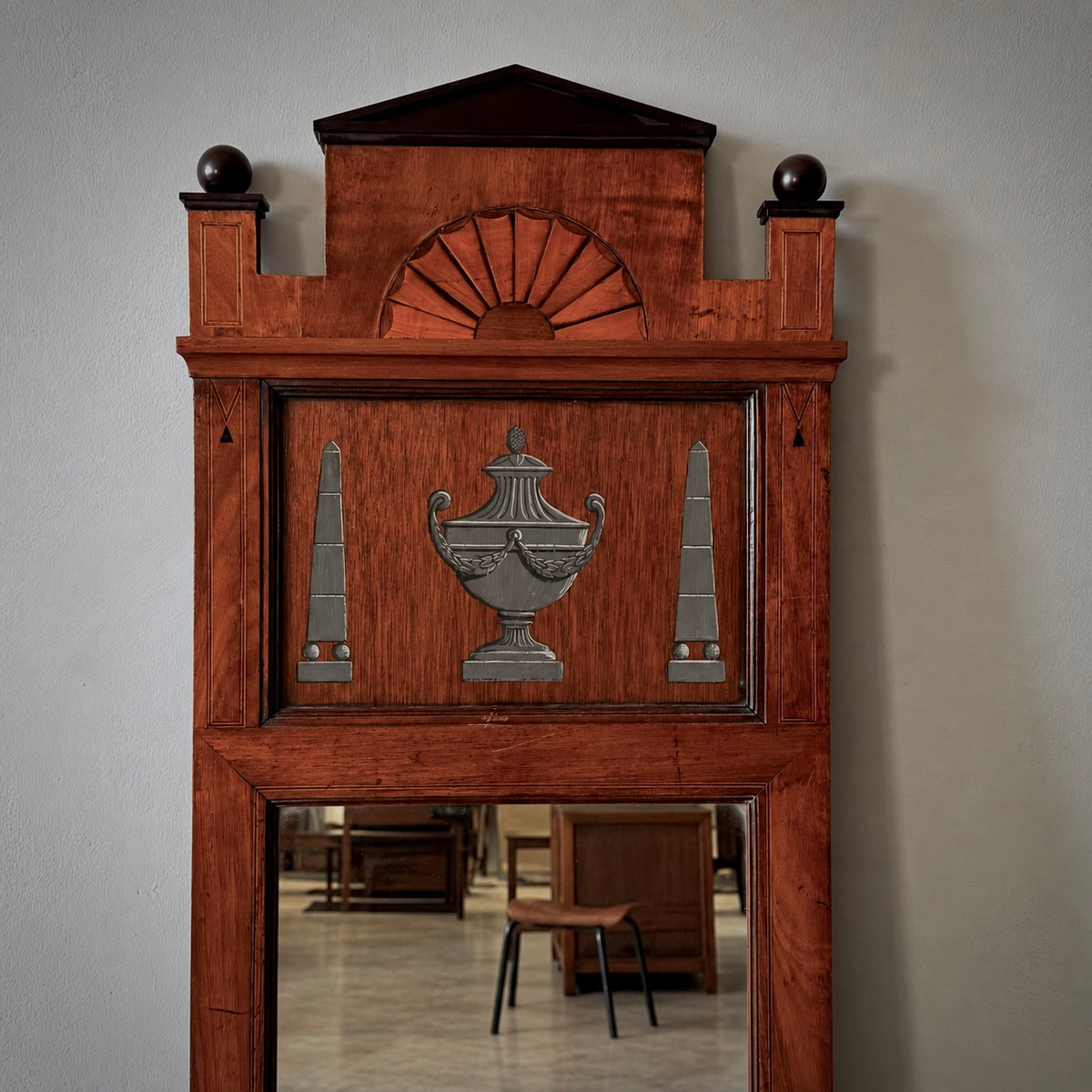 A tall, reddish-brown wooden grandfather clock or wall-hanging piece with decorative metal urn and obelisk motifs on the front, photographed against a plain light gray wall. A mirror at the bottom reflects furniture in an interior room.