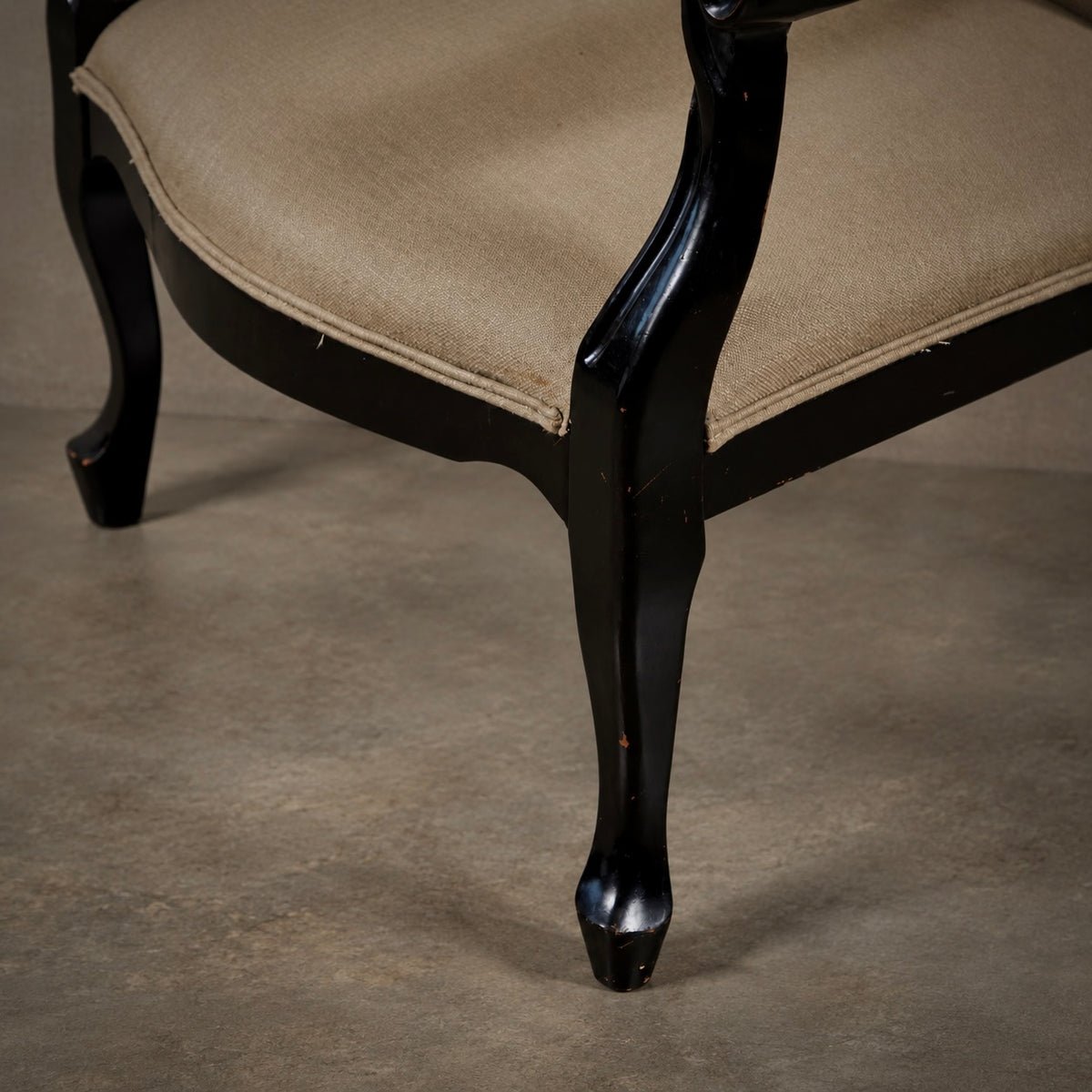 Close-up of a classic armchair featuring a glossy black cabriole-style frame and beige fabric upholstery, shown on a neutral concrete floor.