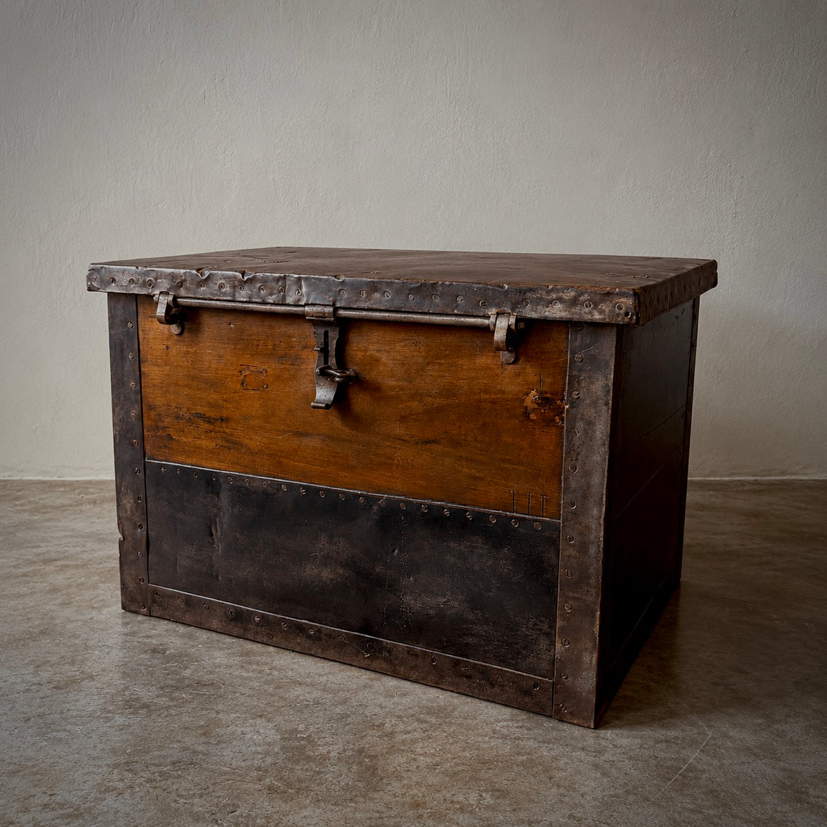 A rustic wooden storage chest with dark metal trim and latch hardware, photographed indoors against a plain light wall and concrete floor.