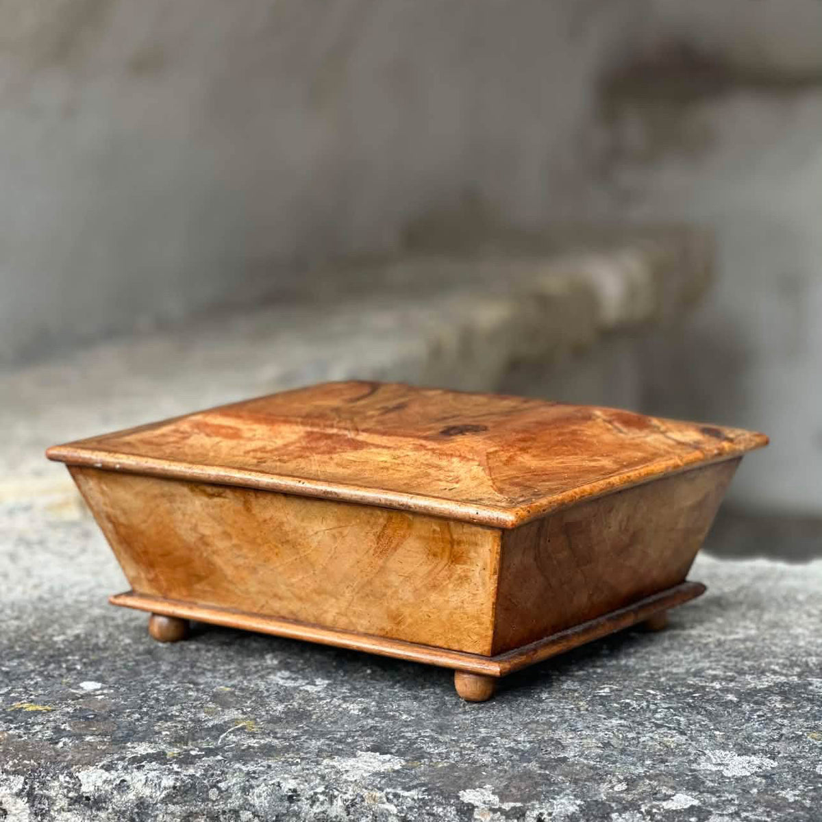 A small square wooden box with a lid, shown on a stone surface against a blurred stone background.