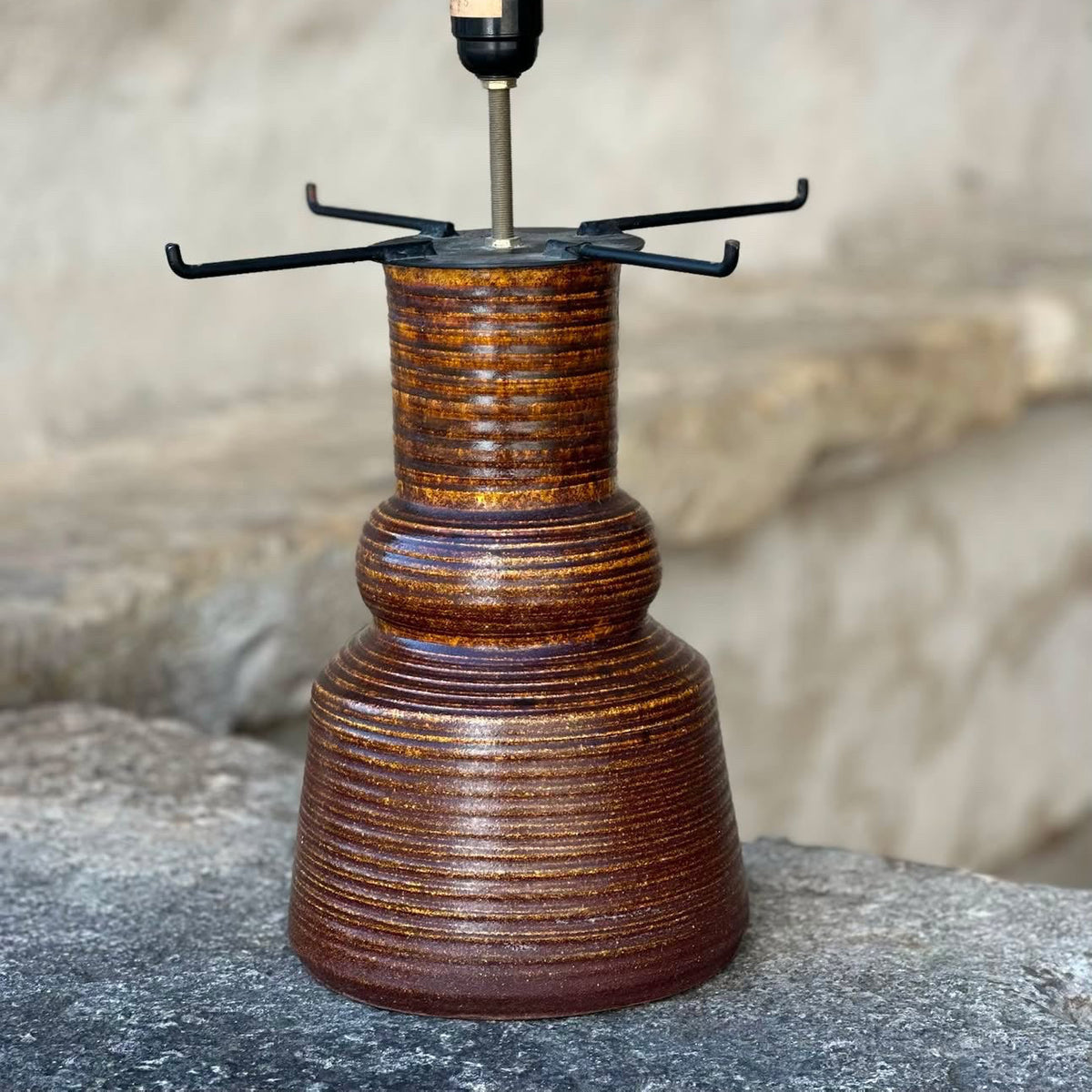 A rustic brown ceramic table lamp with a ribbed textured base, a black metal cross-shaped shade holder, and a clear glass bulb in a black socket. The lamp is displayed outdoors on a stone surface with a blurred stone wall background, and text overlay on the right shows dimensions and price.