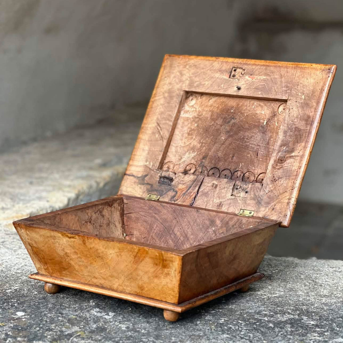 A small carved wooden box with an open hinged lid, shown on a stone surface against a rustic stone wall. The box has a warm honey-brown burl wood finish with darker natural grain patterns.