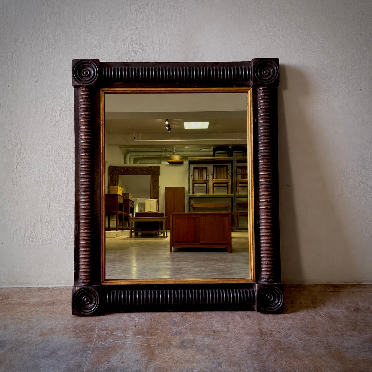 A large rectangular wall mirror with an ornate dark brown carved frame and a gold inner border, leaning against a textured off-white wall on a concrete floor. The mirror reflects an interior room with wooden furniture and shelving.