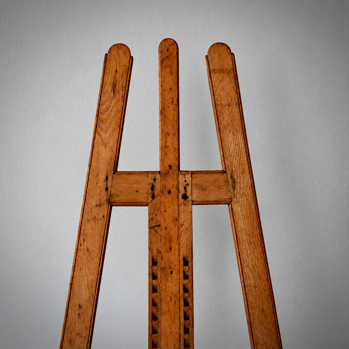 A close-up of a wooden easel or stand with three upright legs and a central crossbar, shown against a plain light gray wall.