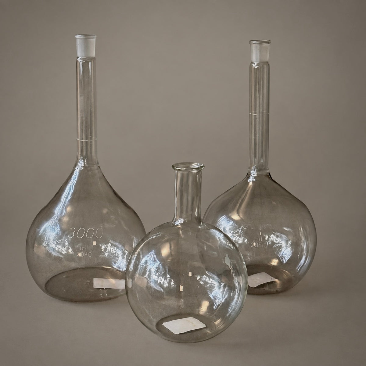 Three clear glass round-bottom flasks with long necks and stoppers, arranged on a neutral taupe background. The front flask is empty, while the two back flasks are partially filled with liquid to demonstrate capacity.