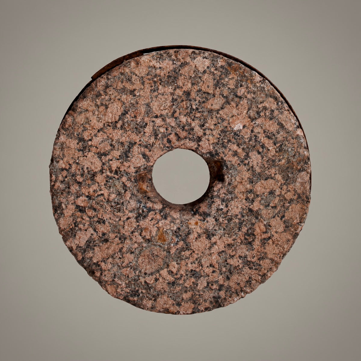 A circular stone or granite ring with a central hole, showing a speckled pink, brown, and black texture, photographed against a solid neutral gray background.