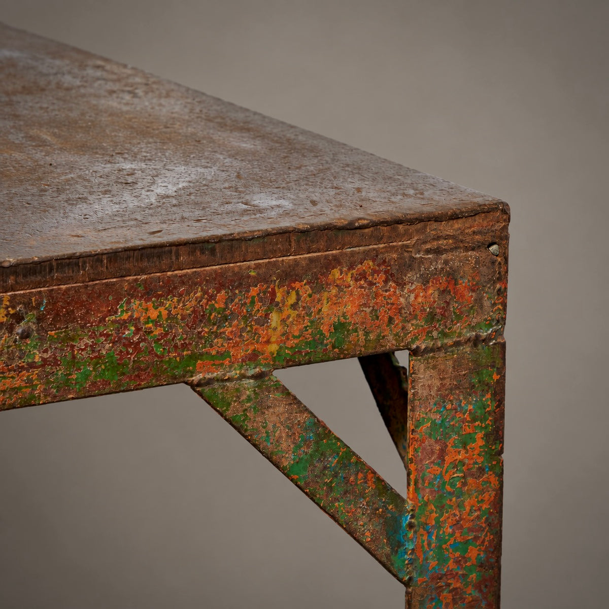 Close-up of a rustic metal table corner showing weathered brown wood and distressed paint with orange and green rust-like patches, against a plain taupe background.