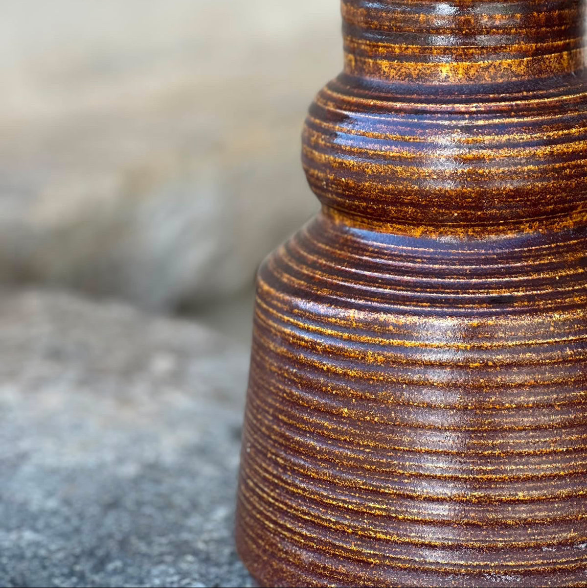 Close-up of a ribbed ceramic vase or container with a glossy brown and amber-orange glazed finish, standing against a softly blurred gray stone background.
