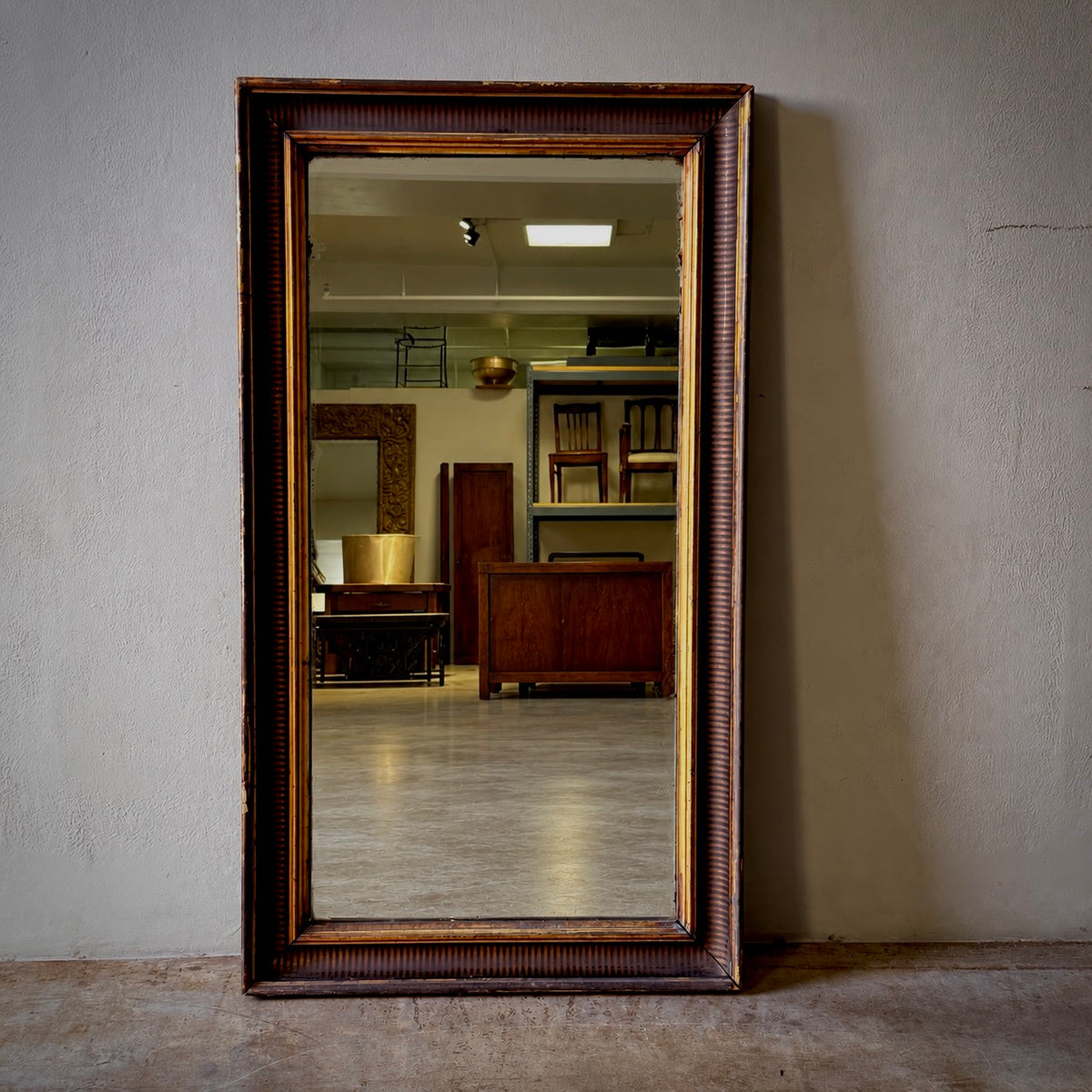 A tall rectangular wooden mirror with a dark brown ribbed frame and gold trim, leaning against a textured beige wall in an interior room.