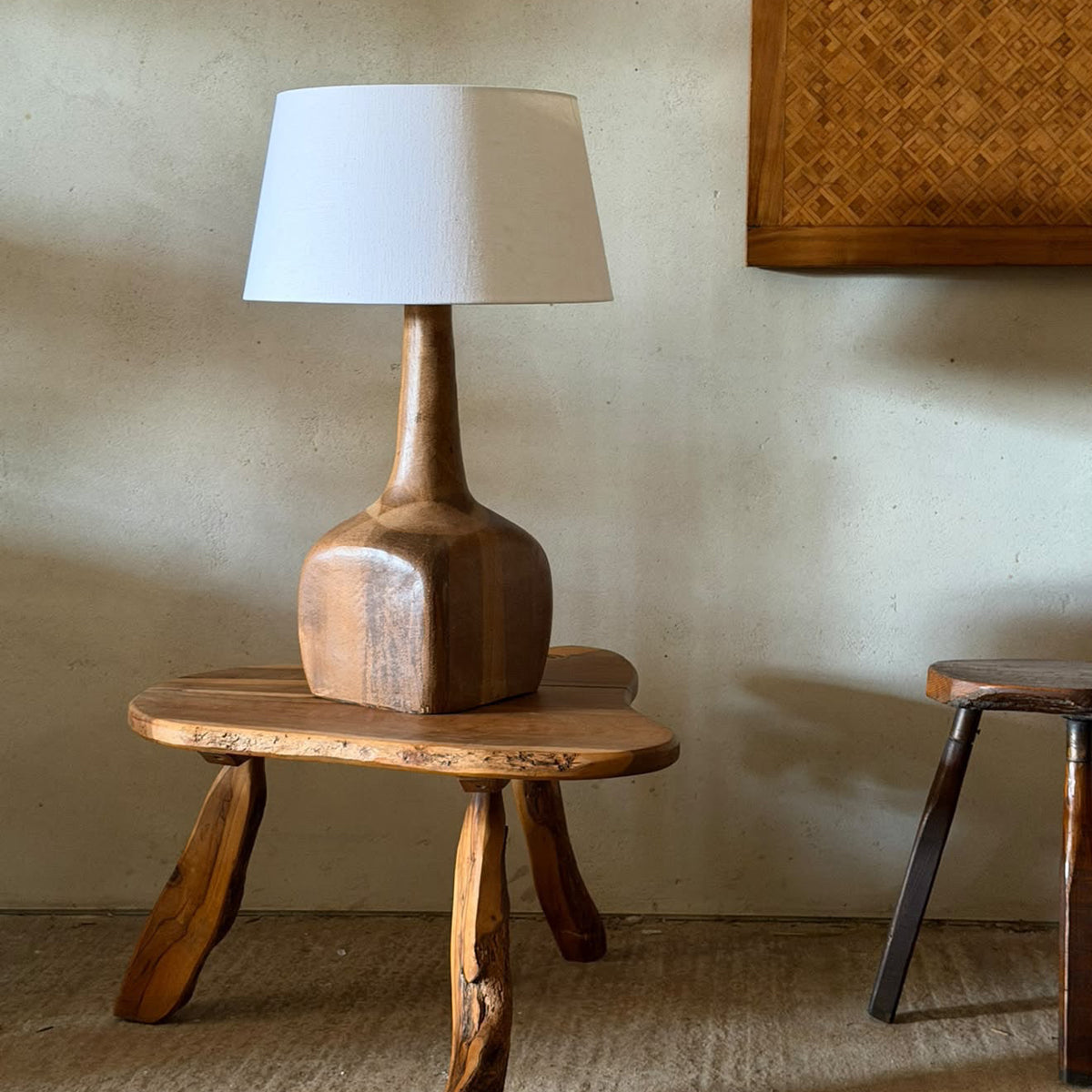 A rustic wooden table lamp with a sculptural, hand-carved oak base and a white drum shade, displayed on a small wooden stool in a warm, neutral-toned room.