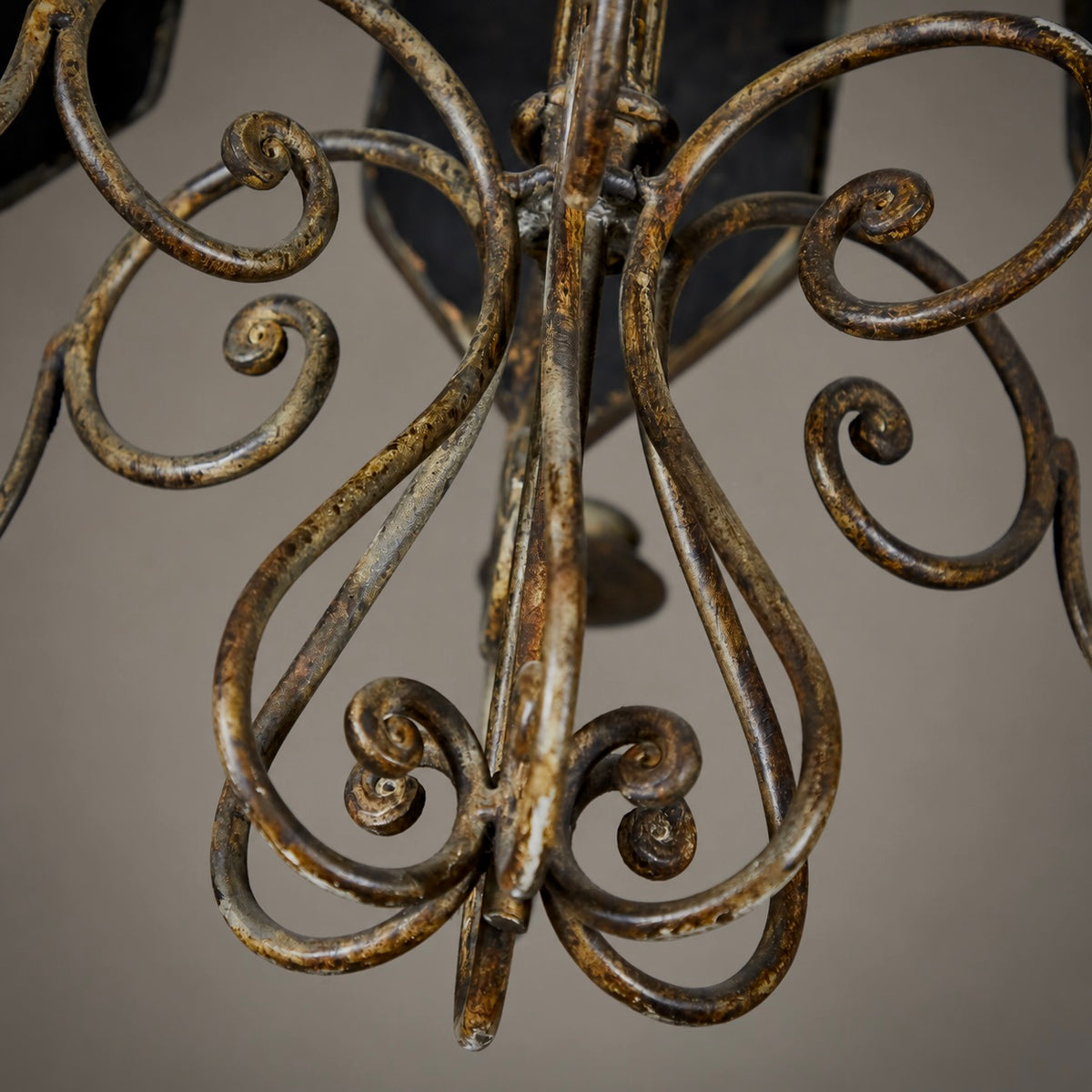 Close-up of an ornate wrought iron chandelier detail, showing scrolled metal arms with a distressed bronze and gold finish against a neutral gray background.