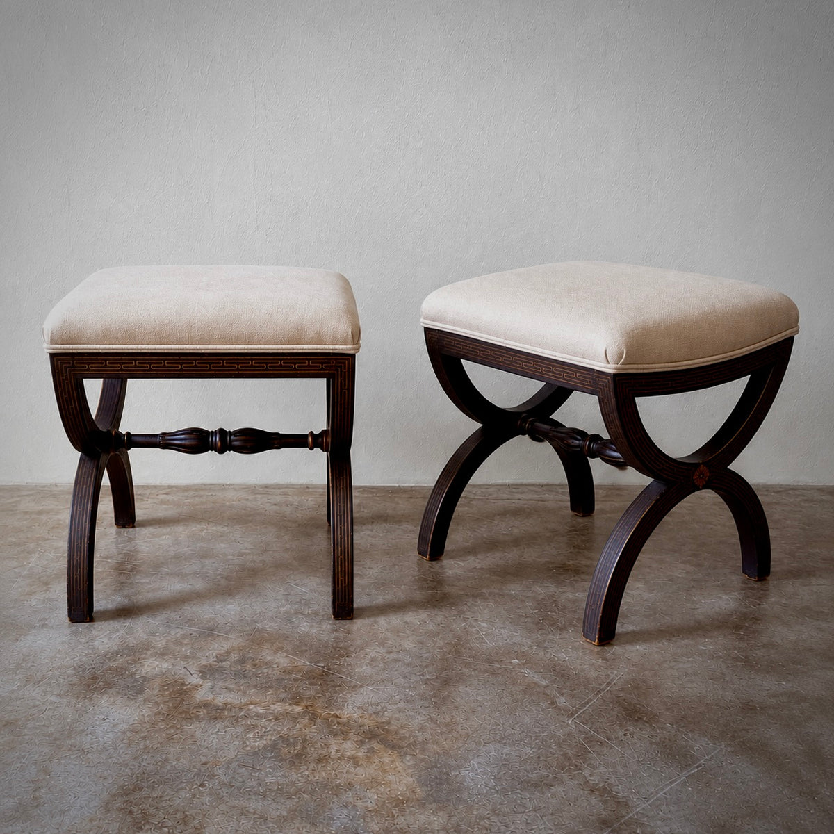 Two wooden stools with cream upholstered square seats and curved dark wood legs, shown on a concrete floor against a textured light gray wall.