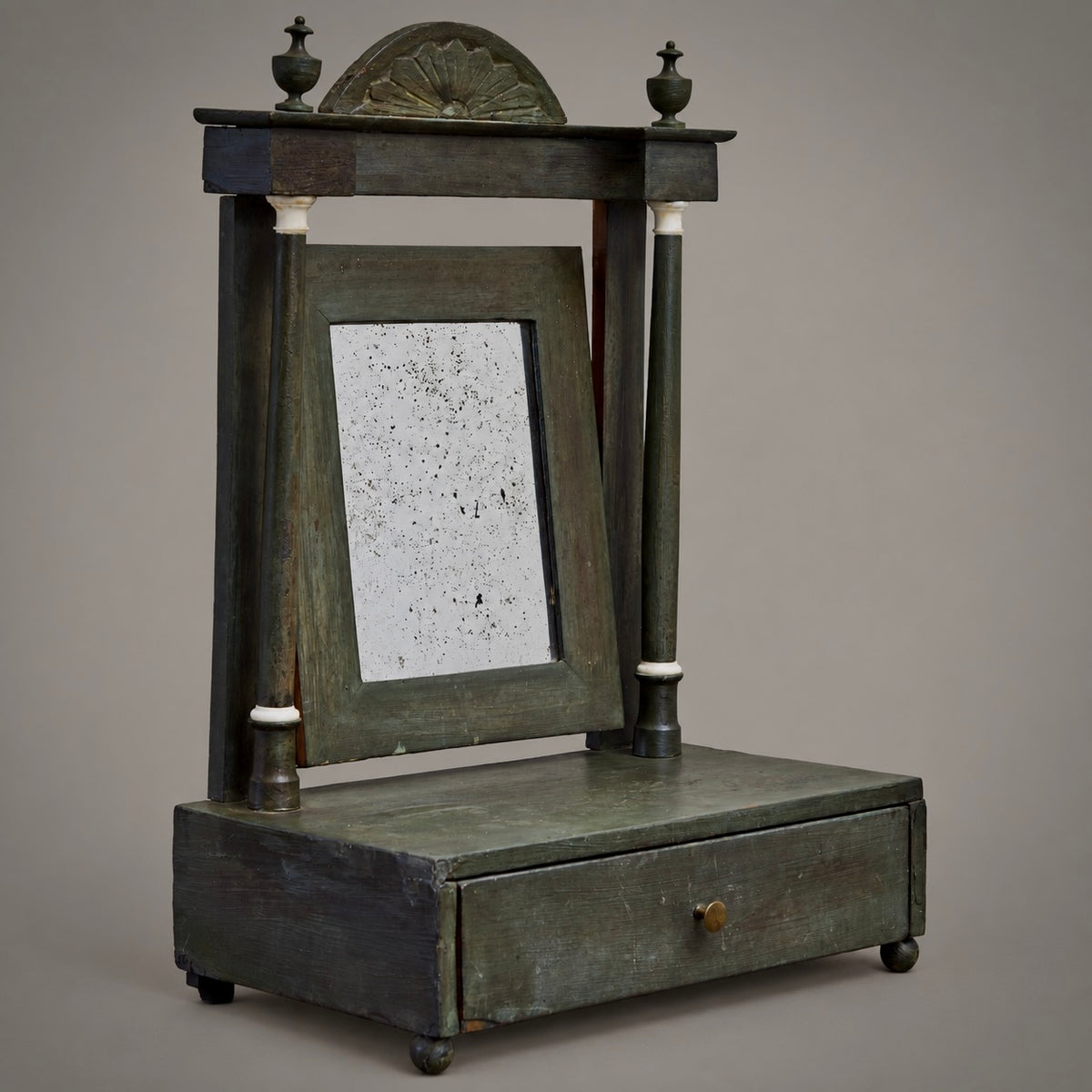 An antique-style wooden cabinet or desk featuring a built-in mirror, a single drawer with a small round brass knob, and decorative finials on top, shown against a neutral gray background.