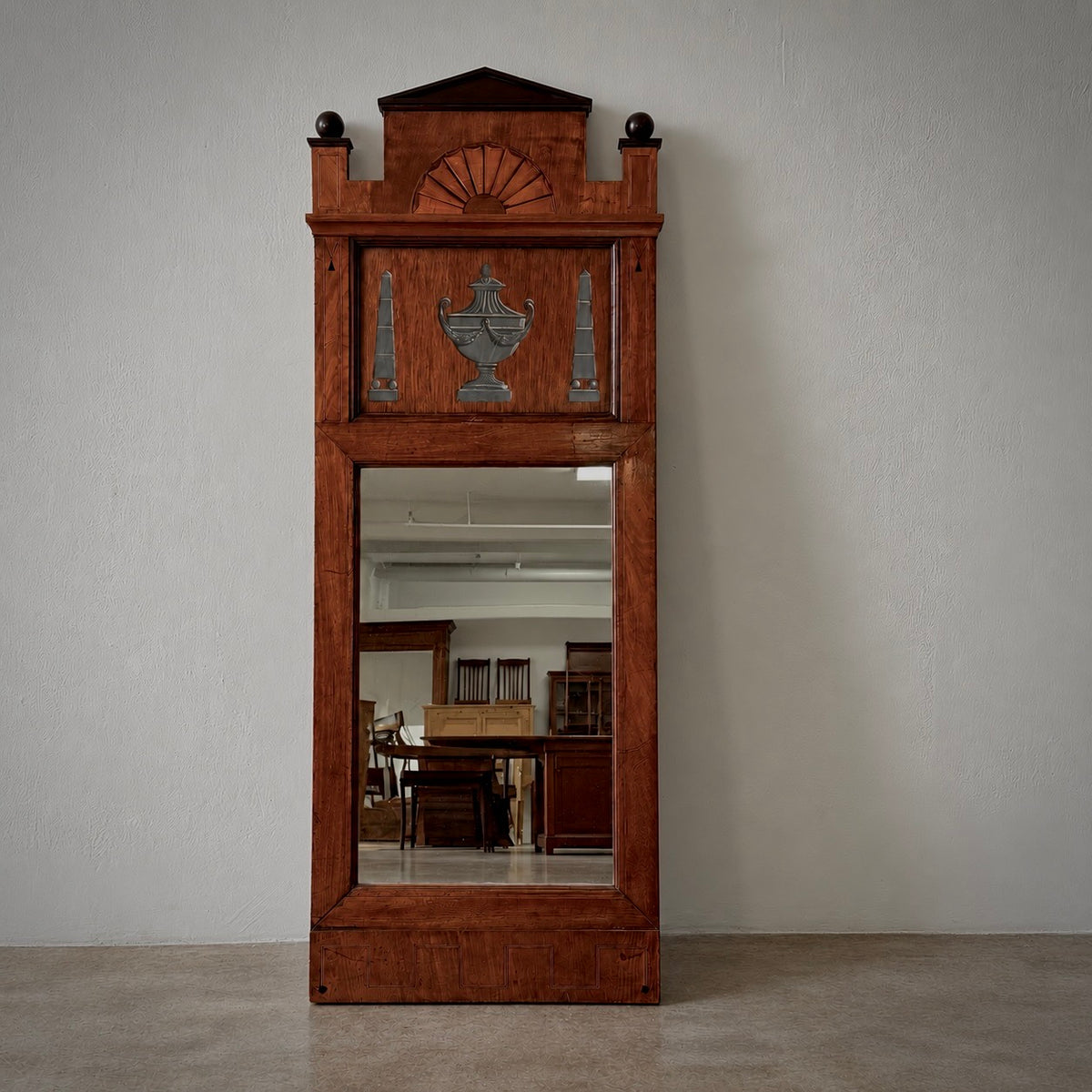 A tall, narrow wooden wall mirror frame with decorative carved details and a large central mirror, set against a plain light gray wall and concrete floor.