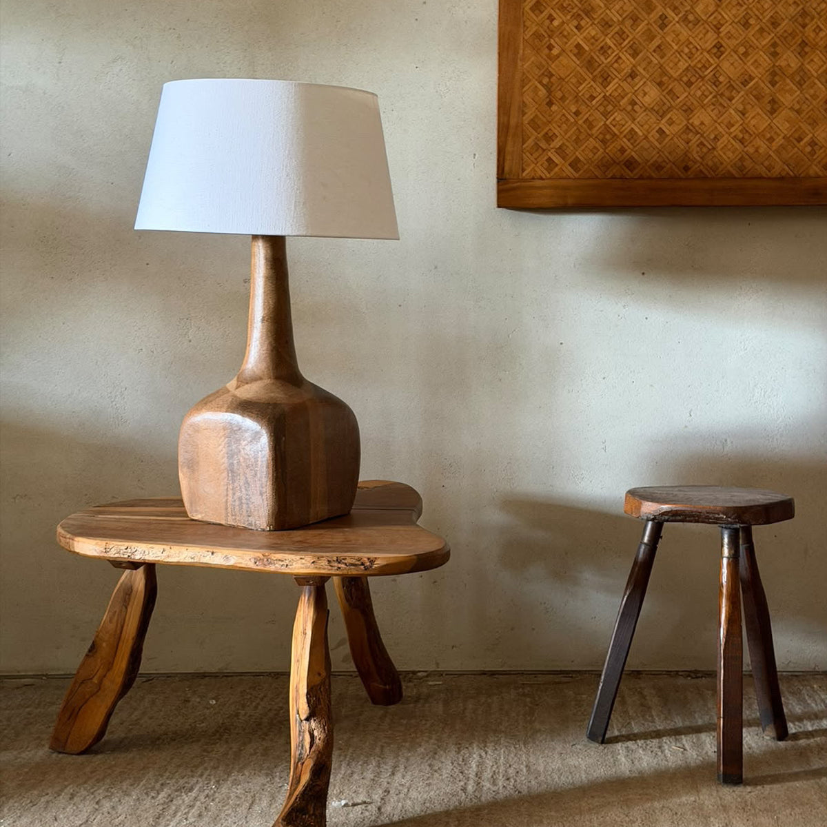 A rustic wooden table lamp with a white drum shade and a carved wooden base, styled on a small wooden stool in a warm, minimalist room.