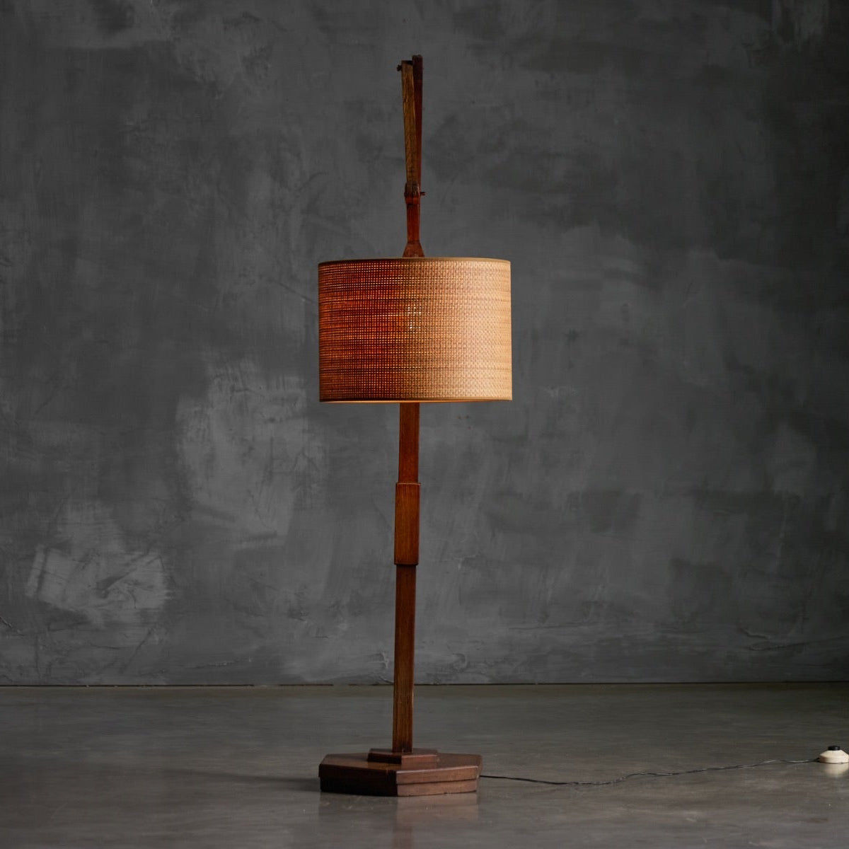 A mid-century modern floor lamp with a cylindrical woven wood shade and a sculptural wooden base, set against a textured gray concrete wall.
