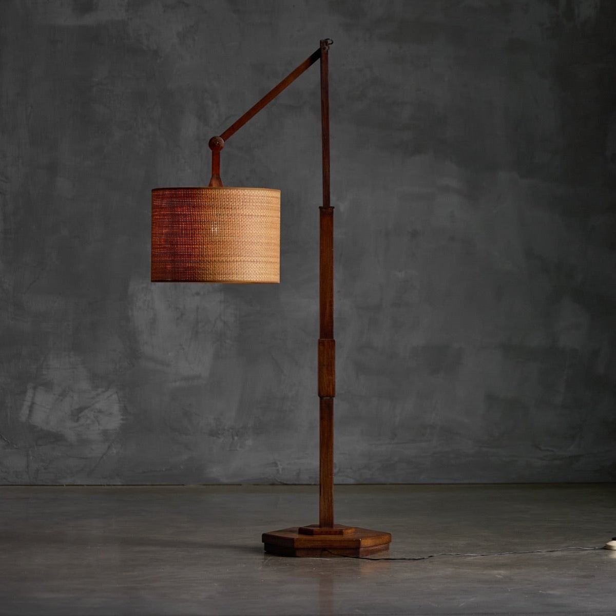 A mid-century modern floor lamp with a dark wood base and adjustable arm, featuring a cylindrical woven bamboo shade, set against a textured gray concrete wall.