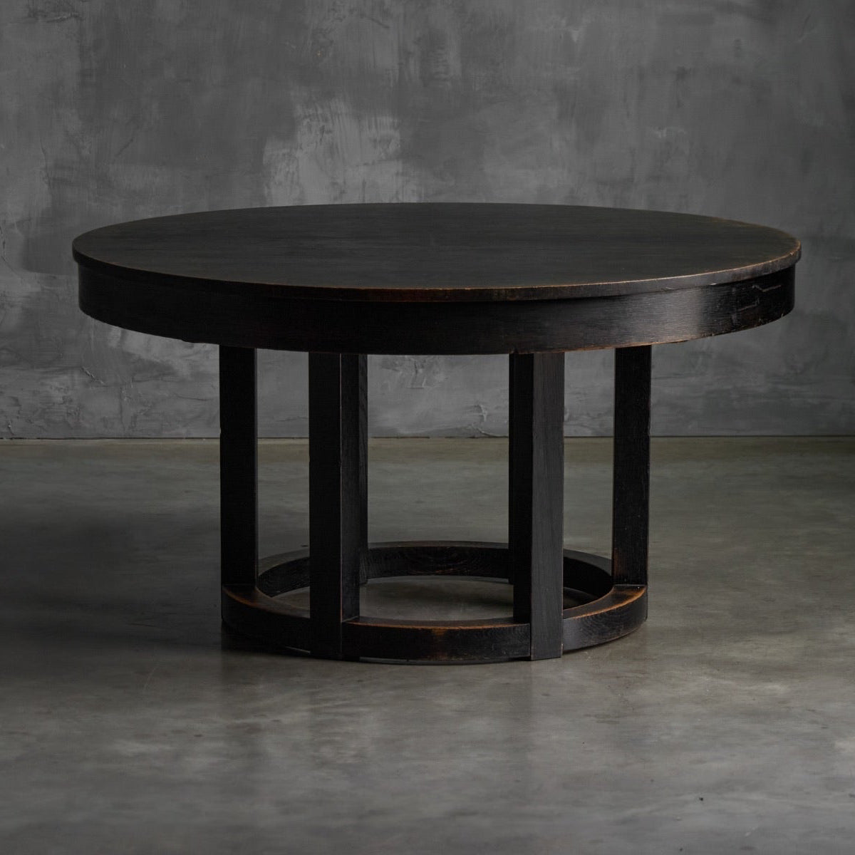 A round dark wood coffee table with a black finish and a multi-tiered geometric pedestal base, set against a textured gray concrete wall and floor.
