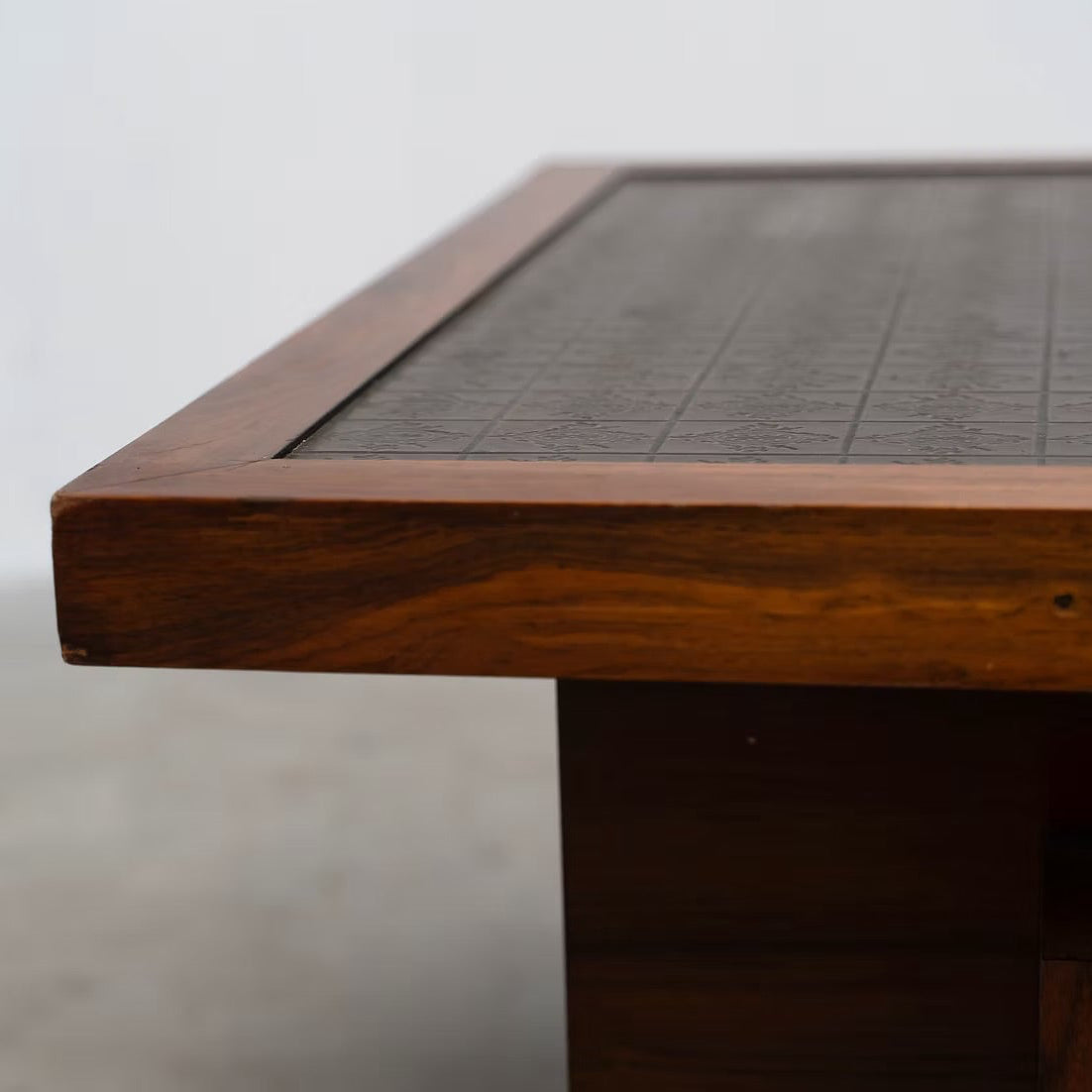Close-up of a wooden table corner with a dark textured tabletop surface and warm medium-brown wood frame, photographed against a plain light background.