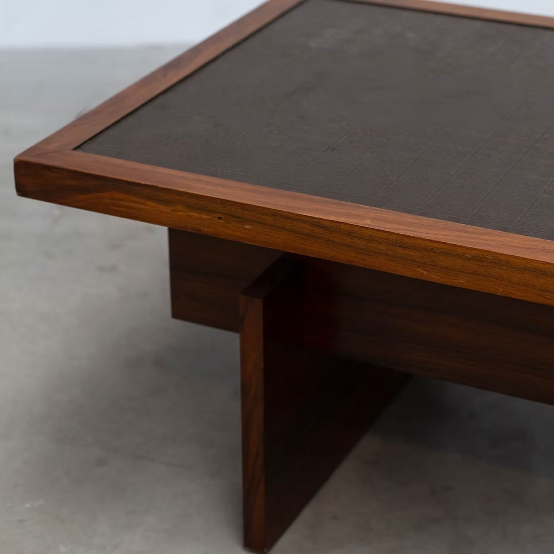 A close-up of a mid-century modern wooden coffee table with a dark brown leather top and a geometric block base, set on a concrete floor.